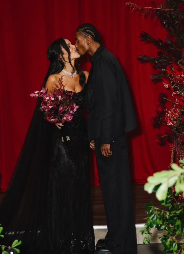 NBA star Kevin Porter Jr. and his wife kiss, both dressed in black, against a red backdrop with red floral arrangements.