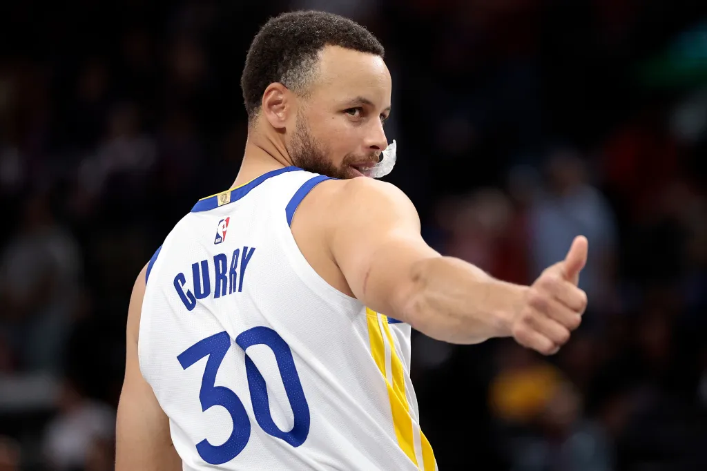 Steph Curry in a white Golden State Warriors jersey gives a thumbs up.