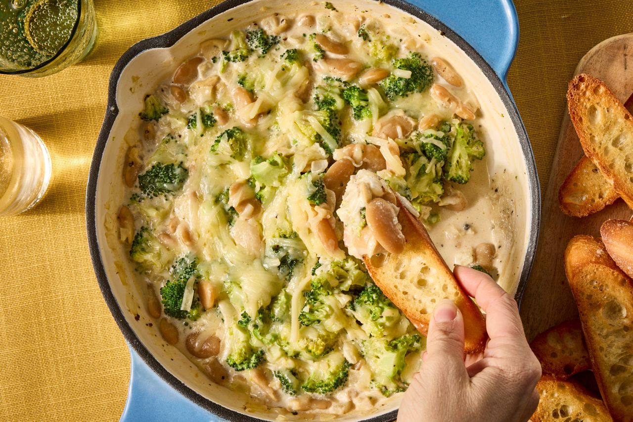A skillet containing a creamy broccoli and bean dip being dipped into with a slice of bread by a hand Credit: Photographer: Jen Causey, Food Stylist: Emily Nabors Hall, Prop Stylist: Hannah Greenwood.