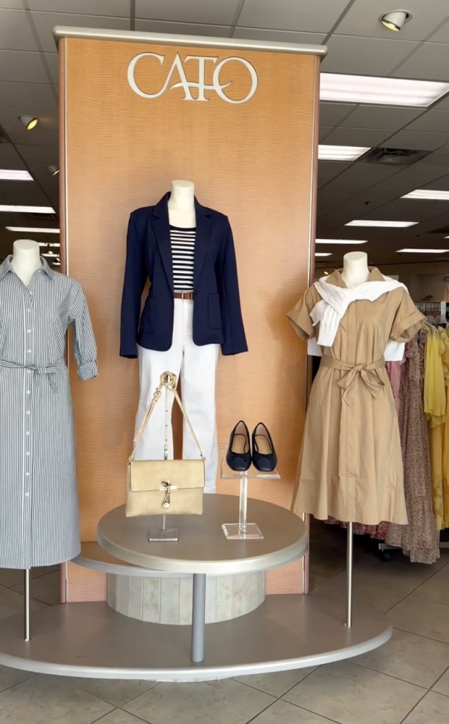 Three mannequins display outfits inside a Cato store, featuring a blue and white striped dress, a blue blazer with white pants and a striped top, and a tan dress with a white sweater.