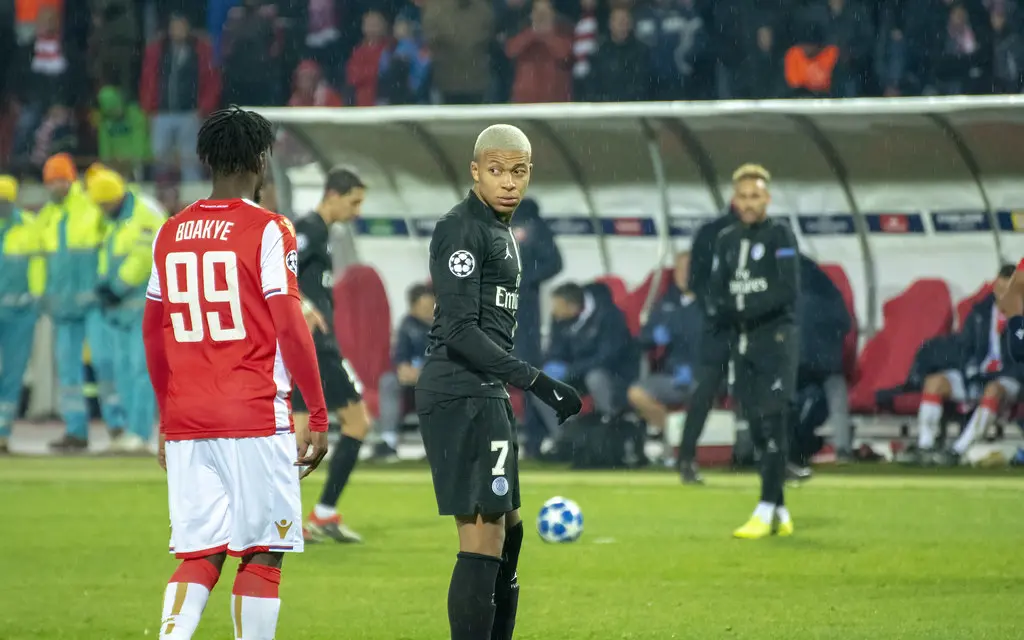 Kylian Mbappé in a black Paris Saint-Germain kit standing on a football pitch with a ball at his feet.