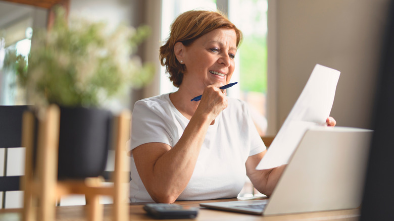A smiling woman sitting in front of an open laptop smiling at a piece of paper