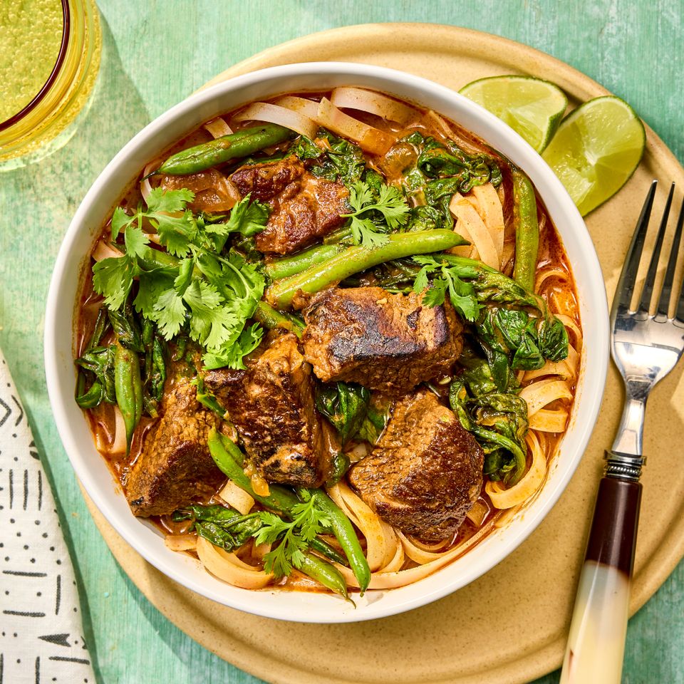 A bowl of beef curry with noodles and vegetables garnished with herbs served with lime wedges and a fork on the side Credit: Photographer: Robby Lozano, Food Stylist: Torie Cox, Prop Stylist: Joshua Hoggle