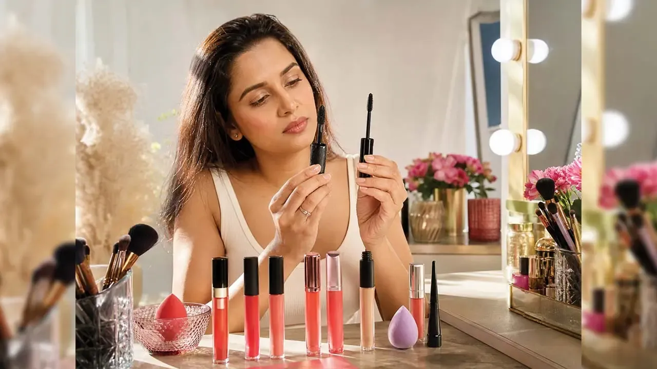 Beautiful Indian woman holding black mascara wand near dressing table with lip glosses, makeup brushes, and vanity mirror.