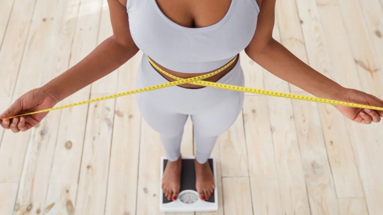 Top-down fitness photograph of a woman measuring her waist with a measuring tape while standing on a weighing scale