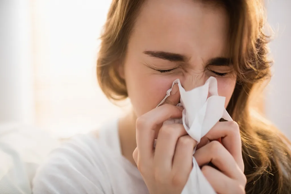 A woman with her eyes closed blows her nose into a tissue.