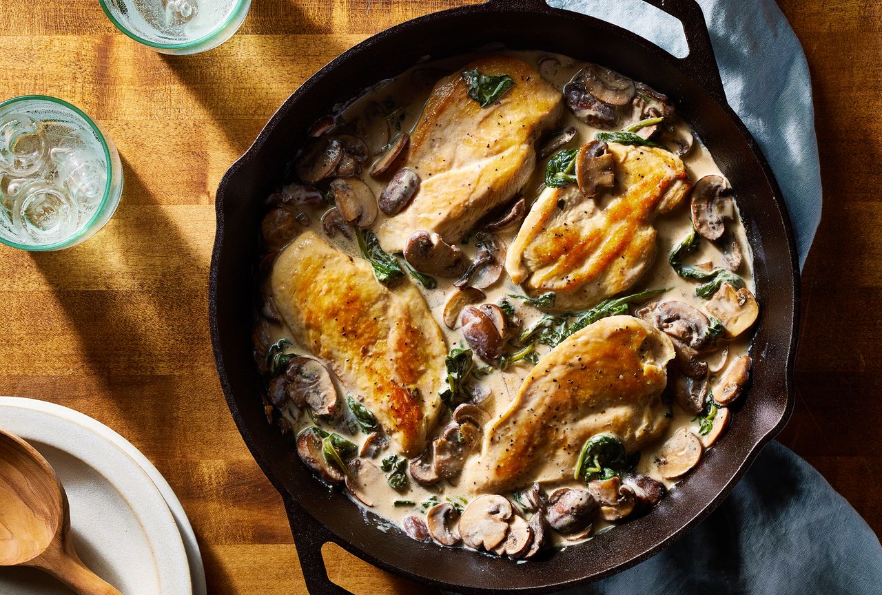 Creamy skillet chicken with spinach & mushrooms recipe, in a cast iron skillet, placed on a wooden surface and a light blue cloth napkin Credit: Photographer: Brie Goldman, Food Stylist: Holly Dreesman, Prop Stylist: Gabriel Greco